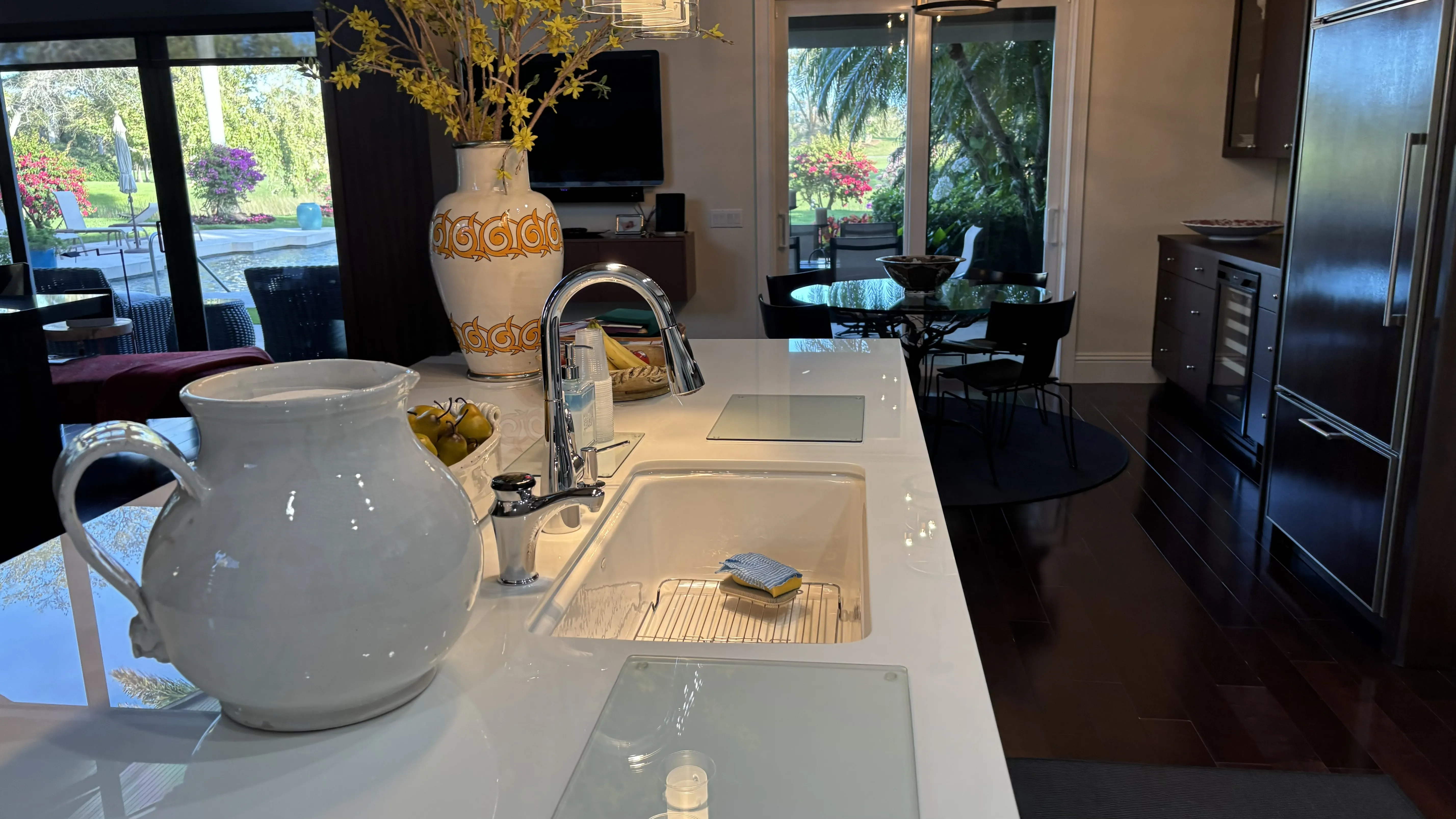 Modern kitchen with a white pitcher, sink, and large windows overlooking a garden.