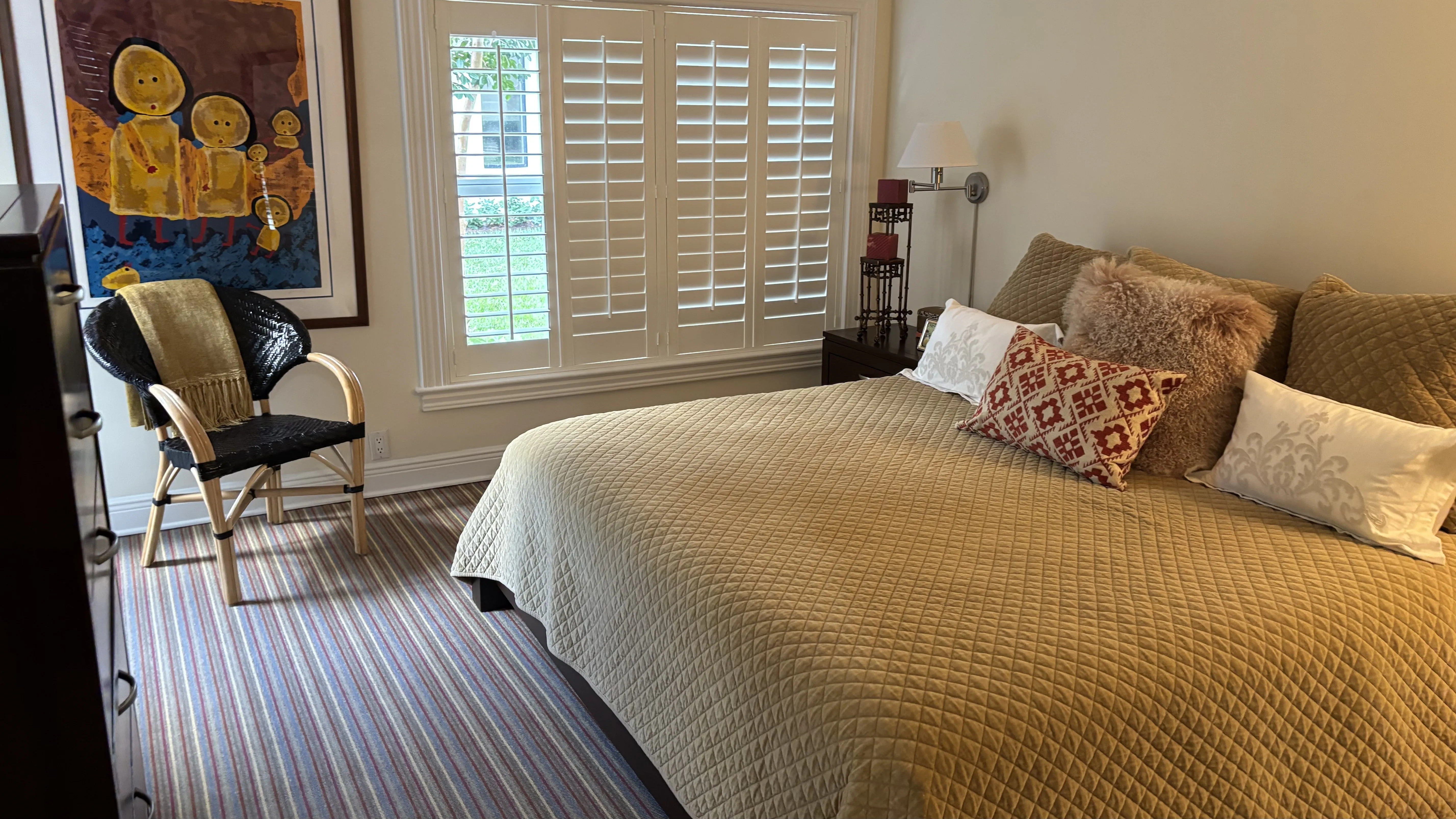 Bedroom with gold quilted bed, varied pillows, striped rug, white shutters, and abstract art.