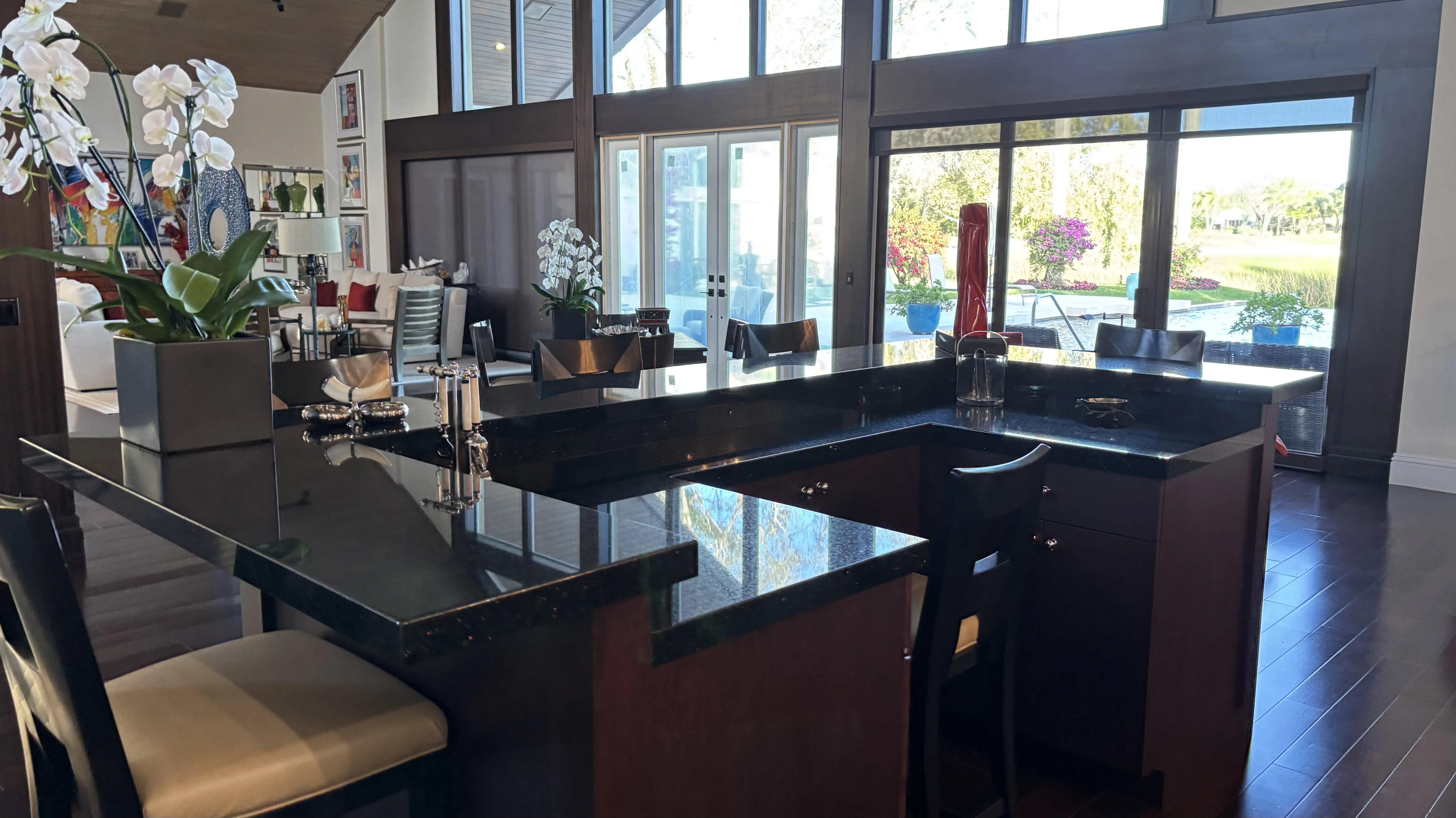 Modern kitchen with dark countertops, bar stools, and large windows overlooking a garden.